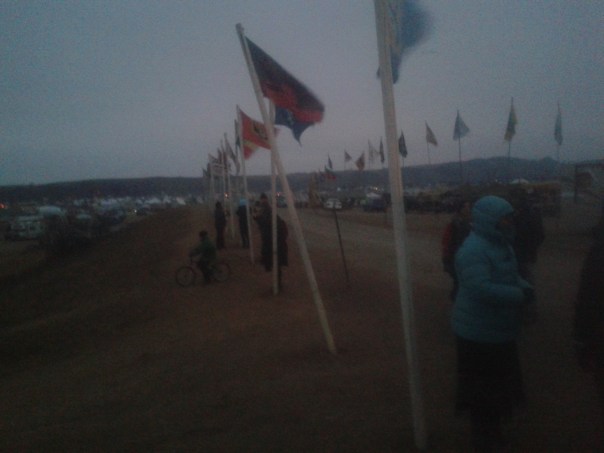 Water protectors are bundled up as the wind blows on Flag Road in the Oceti Sakowin Camp, North Dakota. Flags are billowing, with tents and hills in the background.